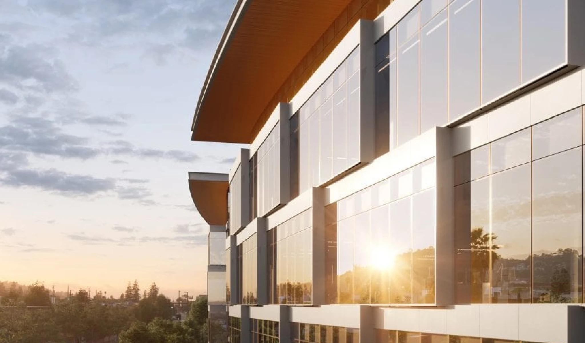 A modern glass and steel building reflects the sunlight during a sunset, with trees and a clear sky in the background.