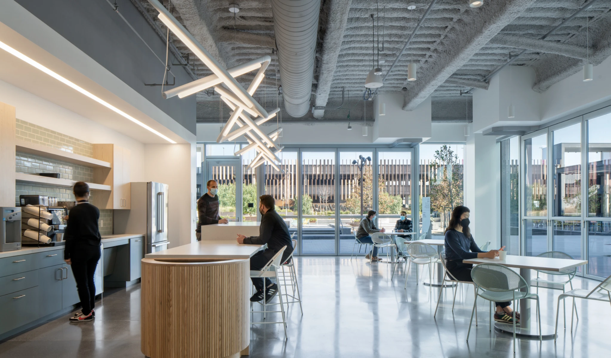 A modern office kitchen and dining area with several people sitting at tables, a person using a coffee machine, and large windows letting in natural light.