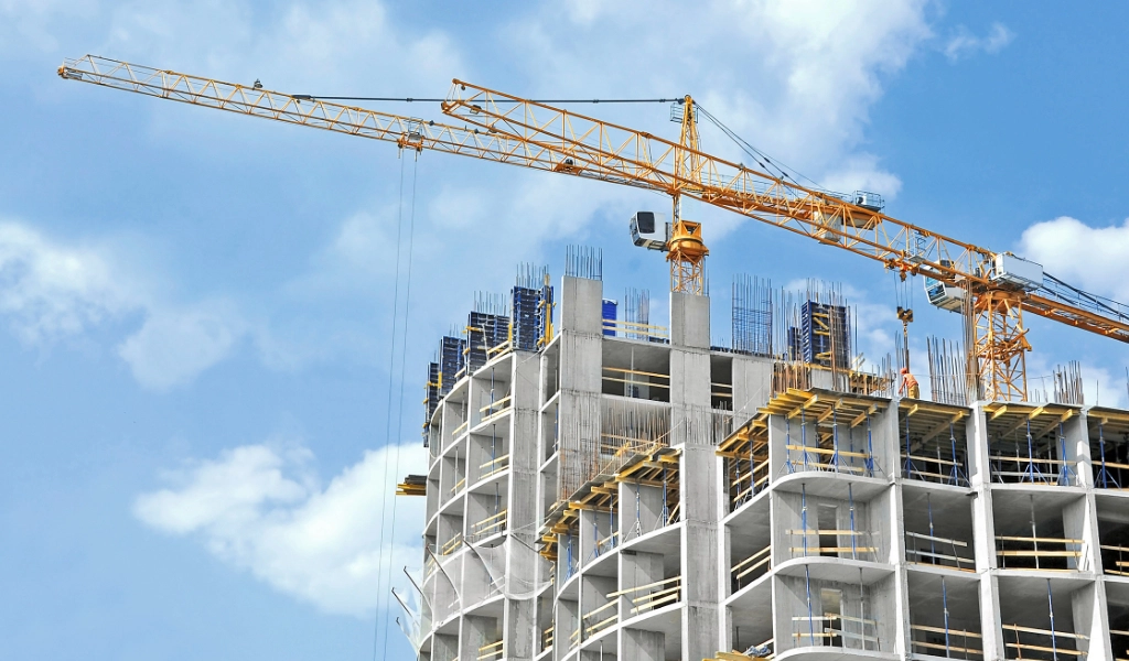 A tall construction crane stands over a multi-story building under construction, with concrete pillars, scaffolding, and steel reinforcements visible against a clear blue sky.