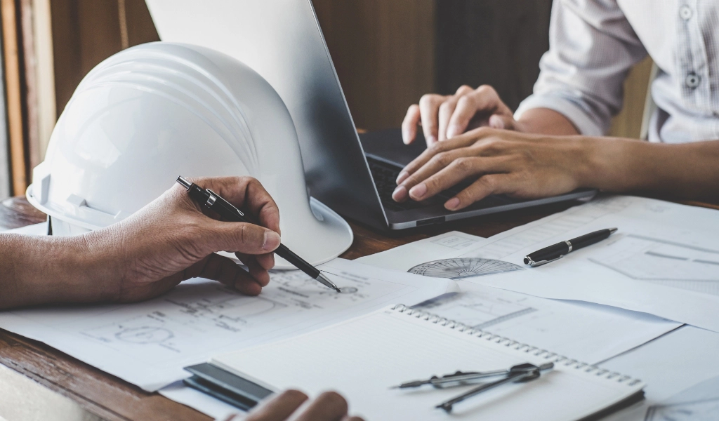 Two individuals working at a desk with construction plans and a laptop. A white hard hat, pencils, and papers are visible on the desk.