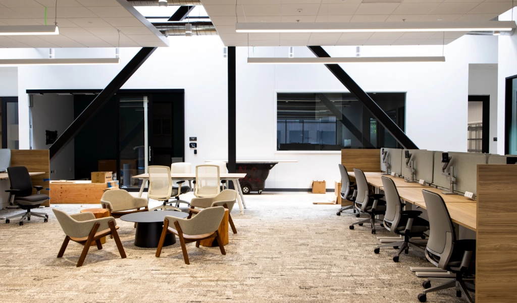 A modern office space with multiple workstations, chairs and desks, a round table with four chairs in the middle, and a few unpacked boxes scattered around. The area is well-lit with natural light.